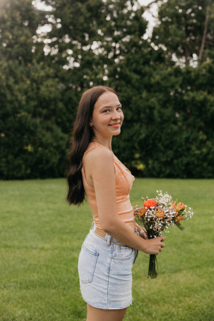 Senior girl in an orange top and denim skirt holding a colorful bouquet of flowers, smiling outdoors during her Minneapolis senior session with Mycah Bain Photography.