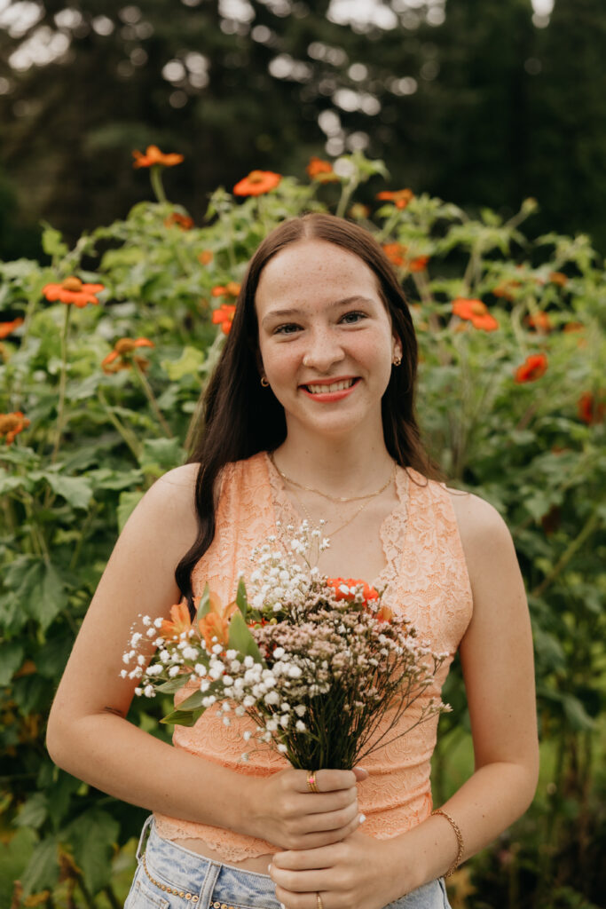 Senior girl smiling while holding a bouquet of orange and white flowers, wearing an orange lace top in a garden with matching blooms, captured by Mycah Bain Photography in Minneapolis.