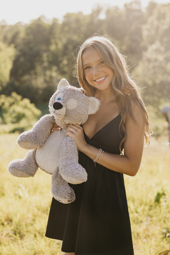 Close-up of a senior girl hugging her teddy bear, glowing in golden hour sunlight while wearing a black dress, captured by Mycah Bain Photography for her Minneapolis senior portraits.