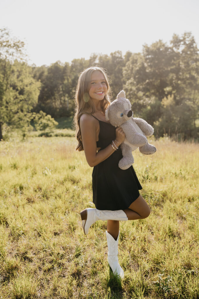 Senior girl in a black dress and white boots holding her childhood teddy bear, smiling in a sunlit field during golden hour senior photos by Mycah Bain Photography in Minneapolis.