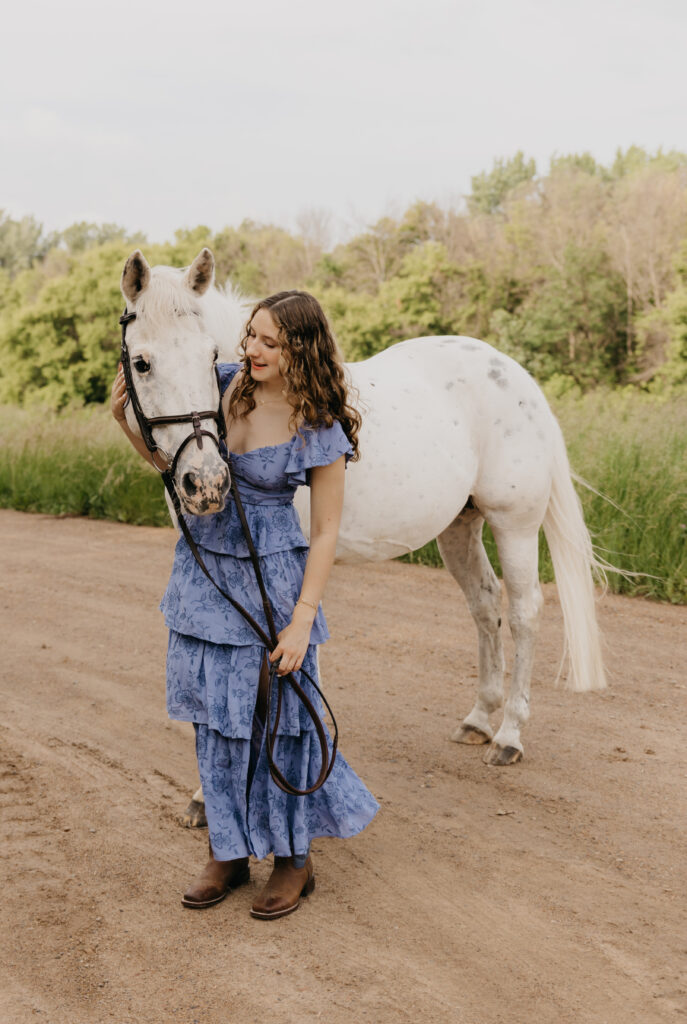 Senior girl in a blue floral dress smiling while walking with her white horse on a dirt road, photographed by Mycah Bain Photography in Minneapolis.