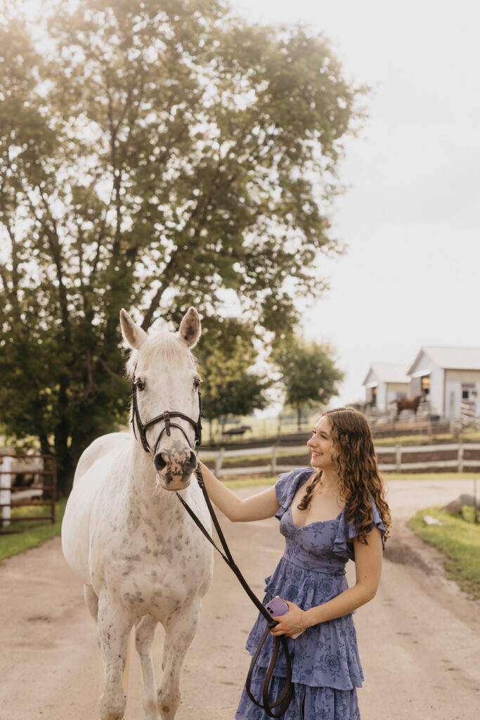 Senior girl looking up at her horse with a smile, holding the reins while wearing a flowy blue dress, captured during her Minneapolis senior photo session by Mycah Bain Photography.