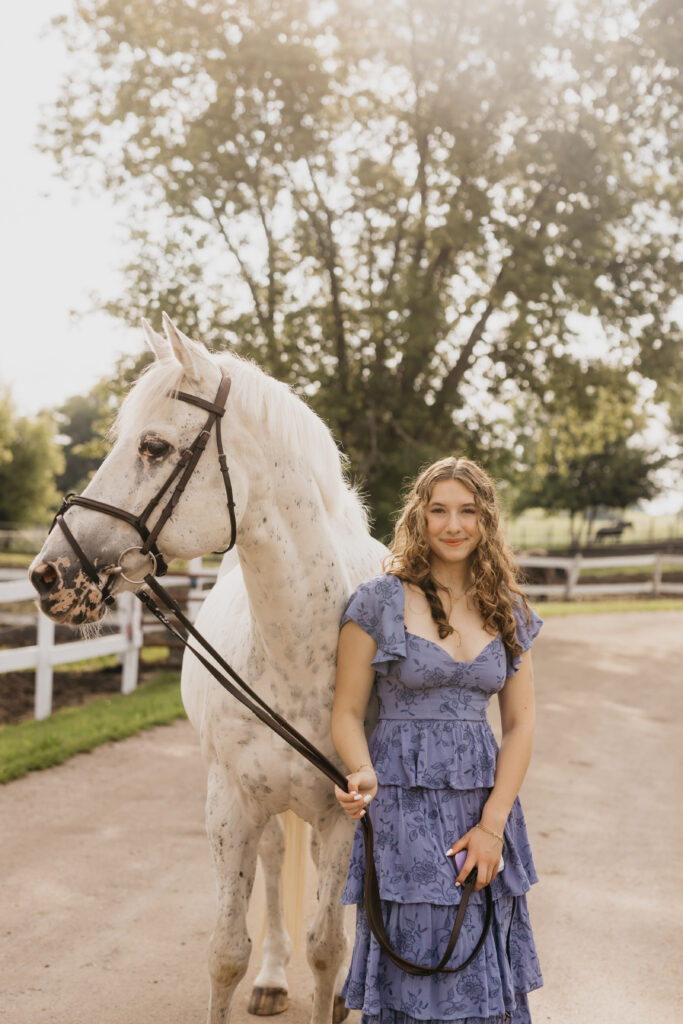 High school senior standing beside her white horse on a farm road, wearing a tiered blue dress during golden hour senior portraits with Mycah Bain Photography.