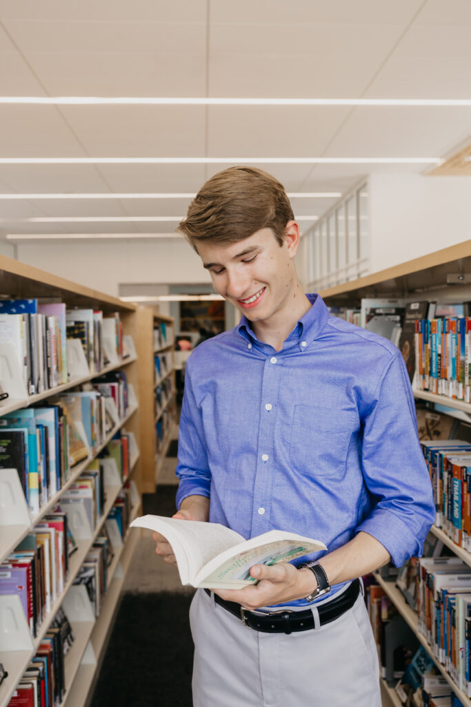 Male student in a blue button-up shirt reading a book while standing between bookshelves in a well-lit library.