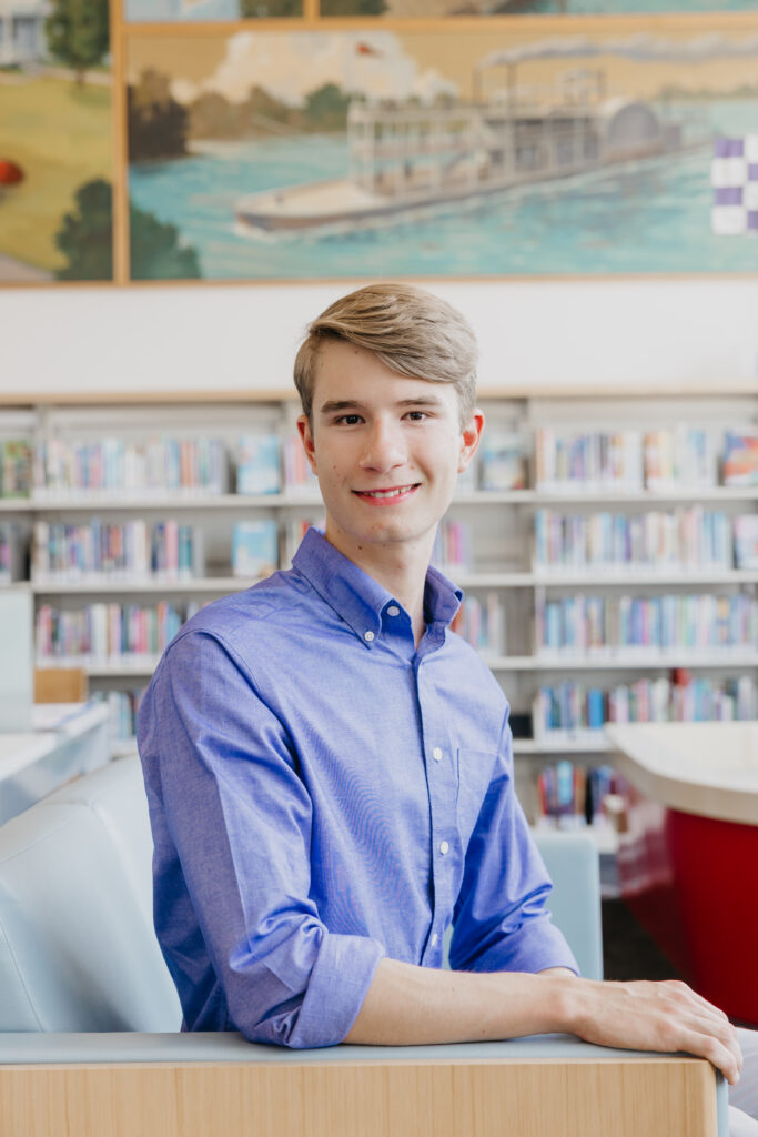 Student in a blue shirt sitting in a library lounge area, smiling at the camera with bookshelves and a mural in the background.