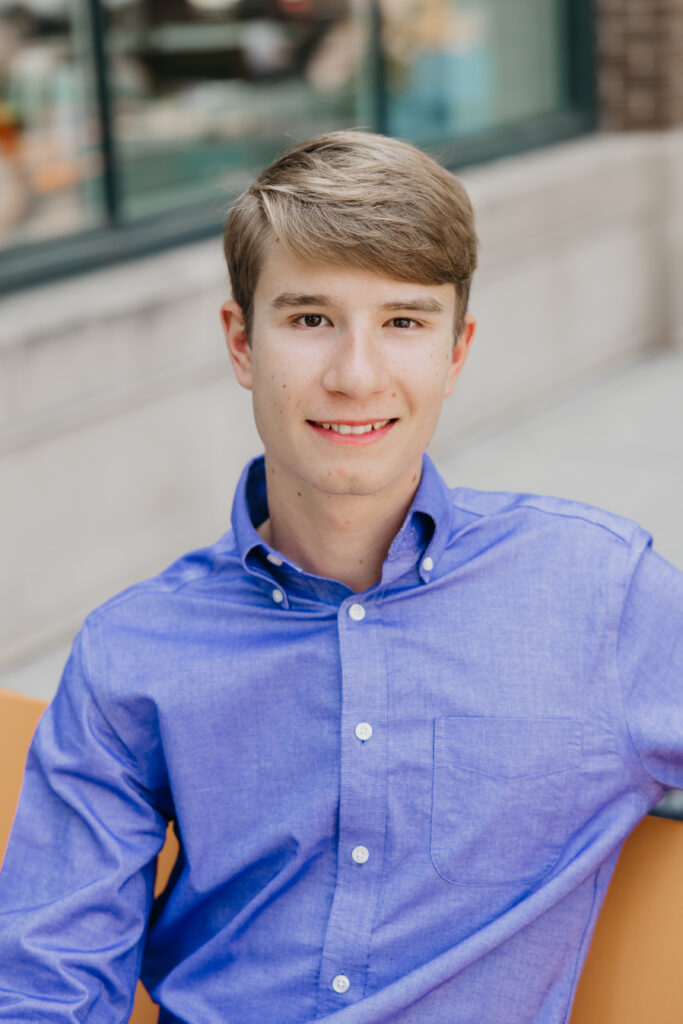 High school senior in a blue button-up shirt sitting on an orange bench in an urban setting, smiling gently at the camera.