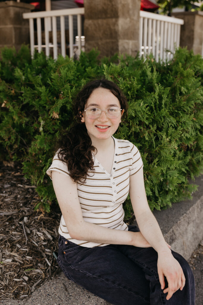 Student sitting on a low stone ledge in front of green bushes, smiling at the camera, wearing a striped shirt and dark jeans.