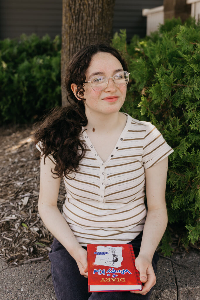 Student with glasses and long dark hair seated near greenery, holding a red Diary of a Wimpy Kid book and smiling softly.