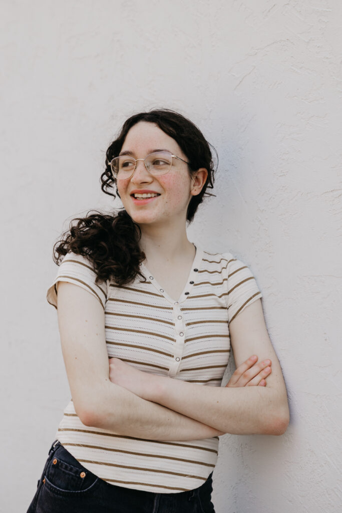 Student with curly dark hair and glasses, smiling while standing against a white wall with arms crossed, wearing a striped short-sleeve shirt and jeans.