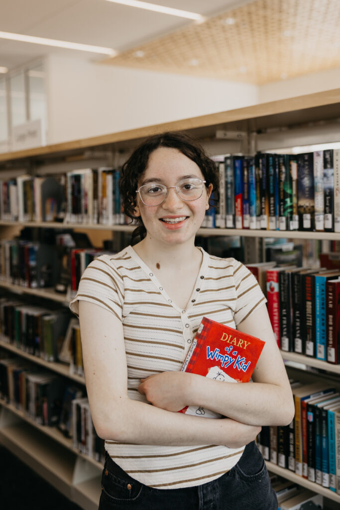 Student standing in front of library bookshelves, holding a copy of Diary of a Wimpy Kid and smiling warmly at the camera.