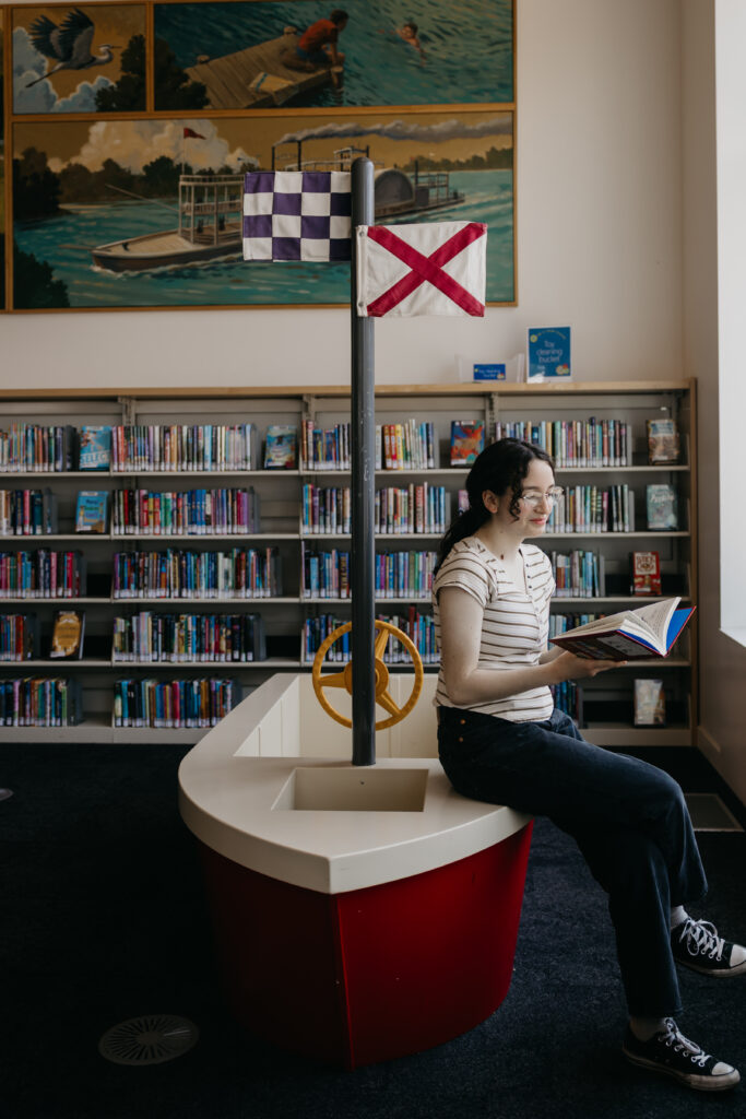 Student sitting on a boat-shaped bench inside a library, reading a book with bookshelves and a nautical mural in the background.