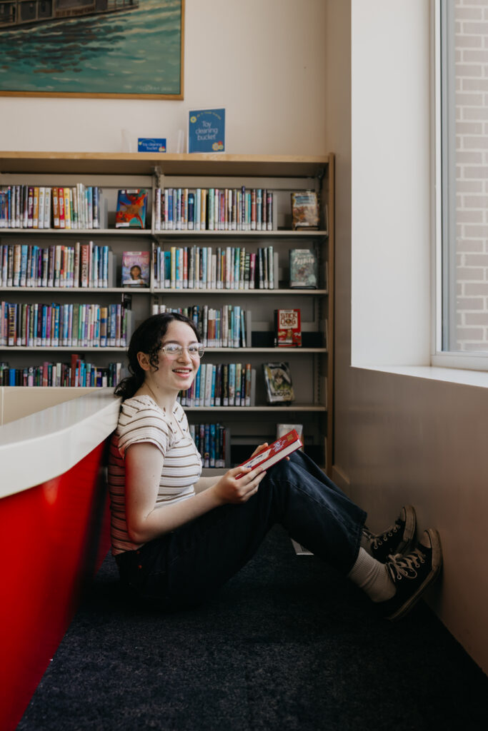Student seated near a large library window, smiling while holding an open red book, with bookshelves in the background.