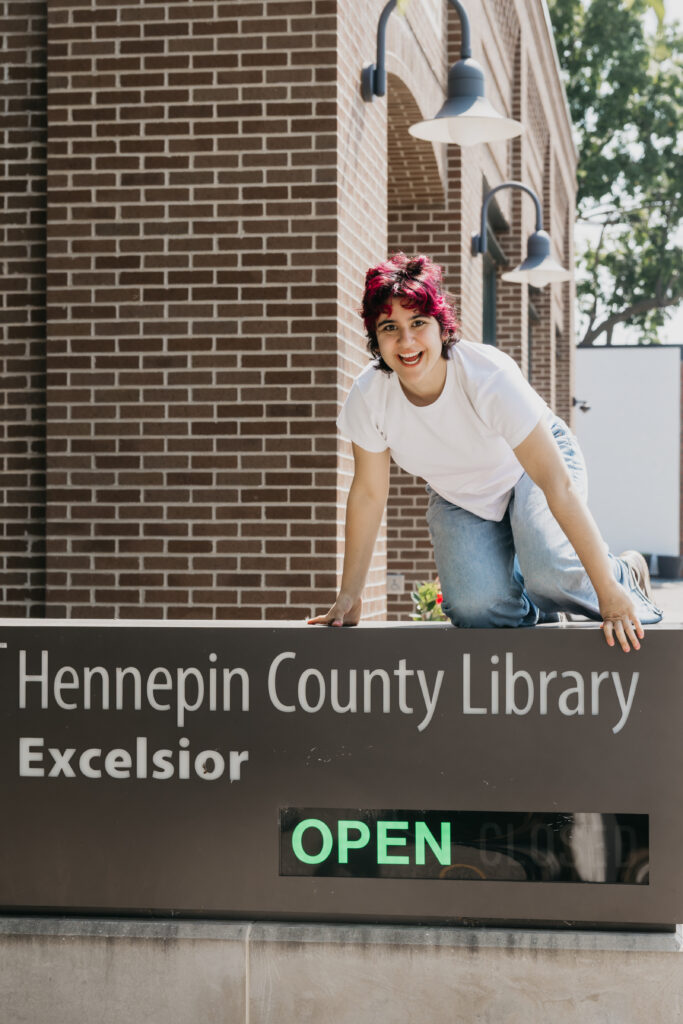 Student in a white t-shirt and jeans playfully posing on top of the Hennepin County Library – Excelsior sign, with a brick building and green 'OPEN' sign visible.