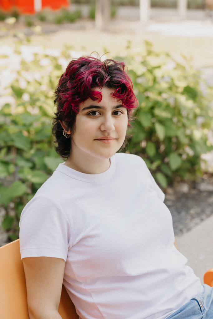 Student with magenta and black hair seated casually on an orange bench outdoors, wearing a white t-shirt and looking at the camera with a gentle expression.