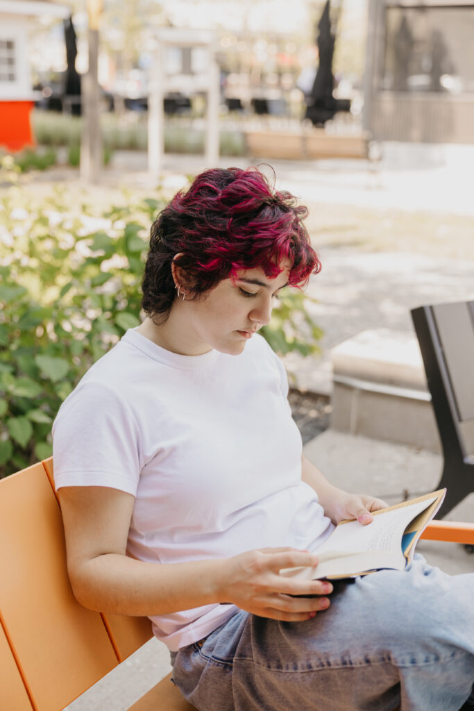 "Student with magenta and black hair reading a book while sitting on an orange bench in a sunny outdoor setting, wearing a white t-shirt and jeans.