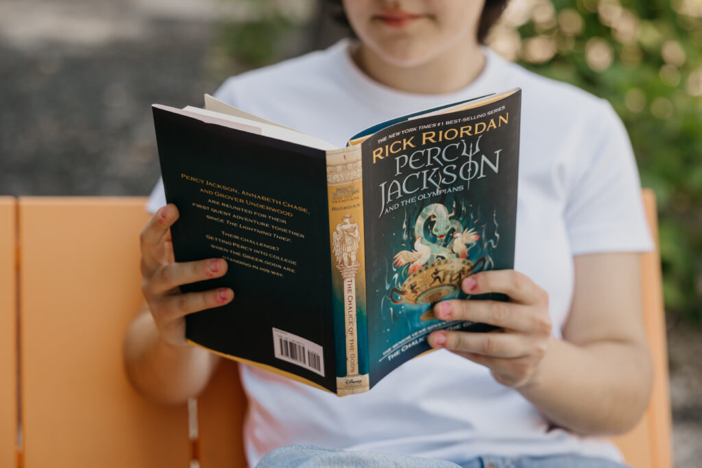 Close-up of a student holding and reading ‘Percy Jackson and the Olympians: The Chalice of the Gods’ by Rick Riordan, wearing a white shirt and jeans.