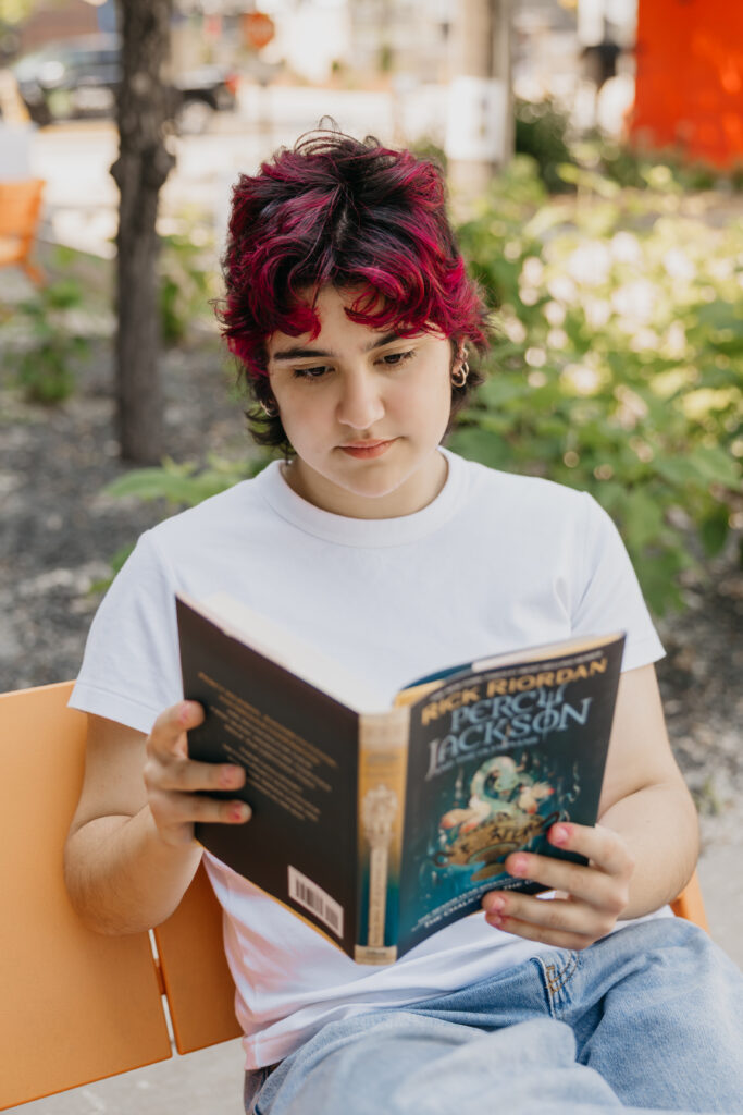 Student with magenta and black hair sitting on an orange bench, reading a Percy Jackson book outdoors, wearing a white t-shirt and jeans.