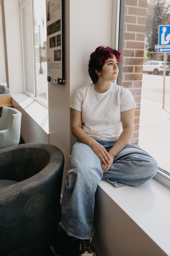 Student with magenta and black hair sitting cross-legged on a windowsill, gazing outside thoughtfully, wearing a white t-shirt and jeans.
