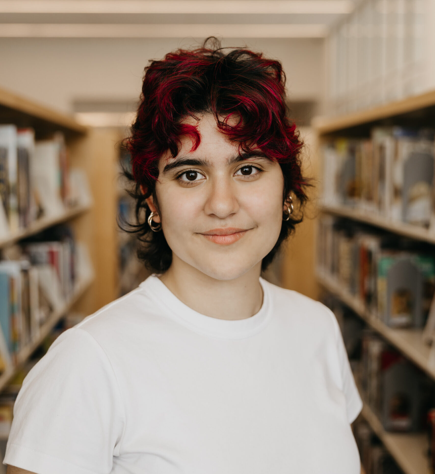 Person with short, curly black hair with vibrant magenta highlights, wearing a white t-shirt, standing outside in front of a brick wall and window, looking confidently at the camera with a subtle smile.