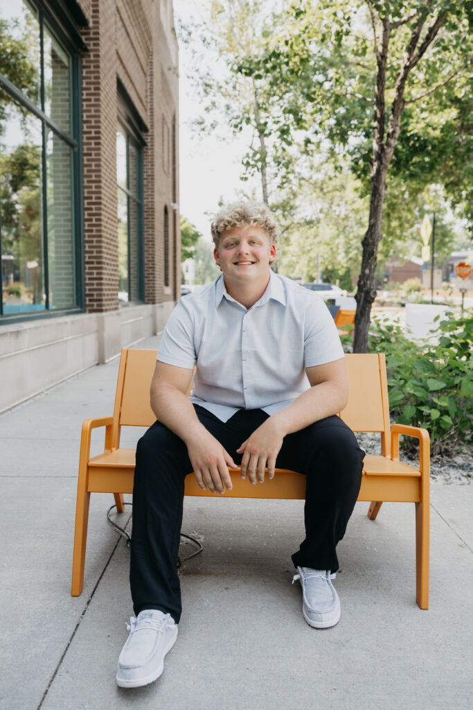 Student sitting on an orange bench outside a brick building, smiling with hands resting on his knees, surrounded by trees and sidewalk greenery.
