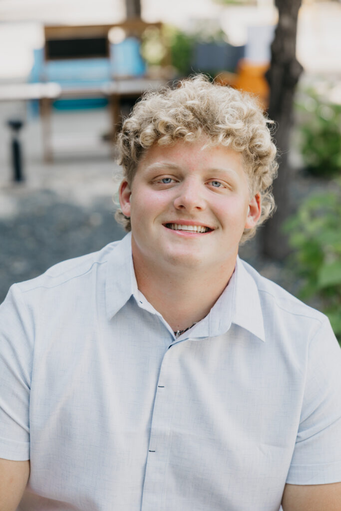 Blond male student smiling directly at the camera in an outdoor setting, wearing a light blue collared shirt.