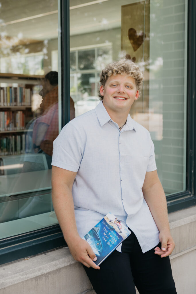 Male student with curly blond hair leaning casually against a window outside the library, holding The Young Man and the Sea and smiling.