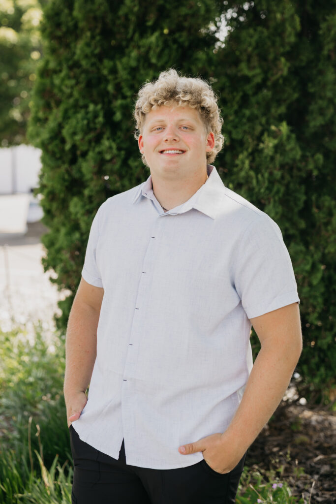 Male student with curly blond hair standing in front of a tall green tree, smiling with hands in his pockets, wearing a light blue collared shirt.