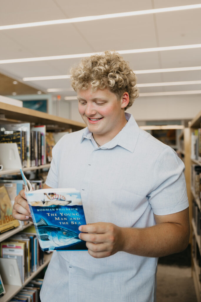 Student with curly blond hair wearing a light blue shirt, smiling while reading The Young Man and the Sea in a library aisle.