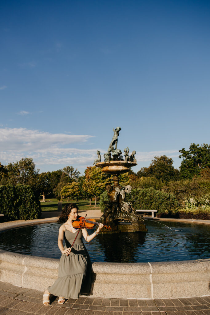 Senior girl playing the violin in front of a large fountain at a Minneapolis park, captured during golden hour senior portraits by Mycah Bain Photography.