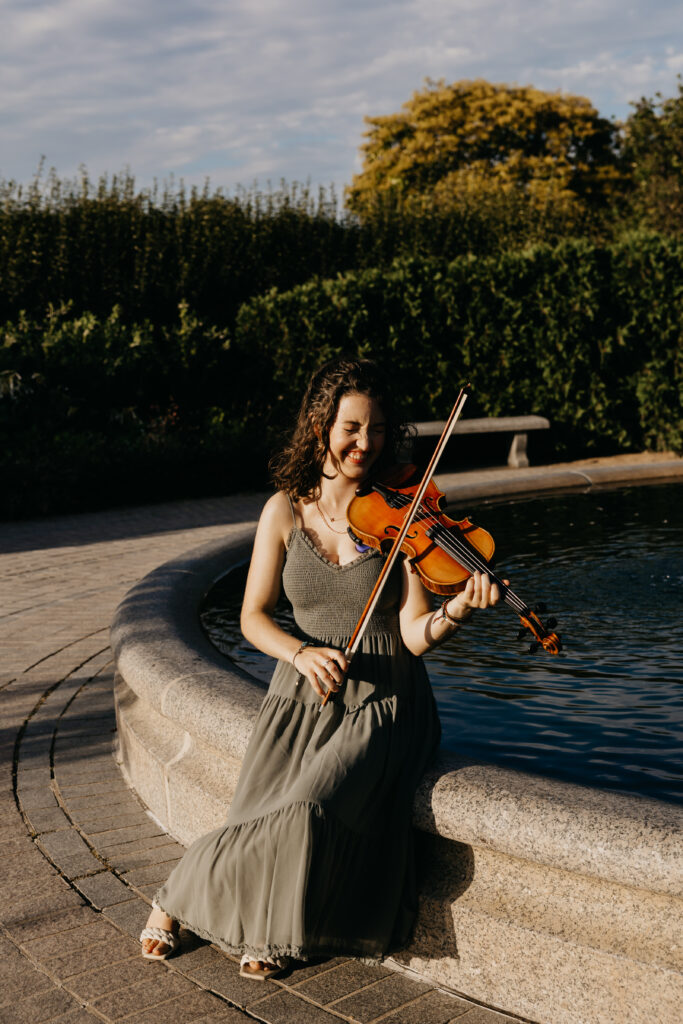 High school senior smiling while playing the violin next to a stone fountain, wearing a gray dress during her Minneapolis senior photo session with Mycah Bain Photography.