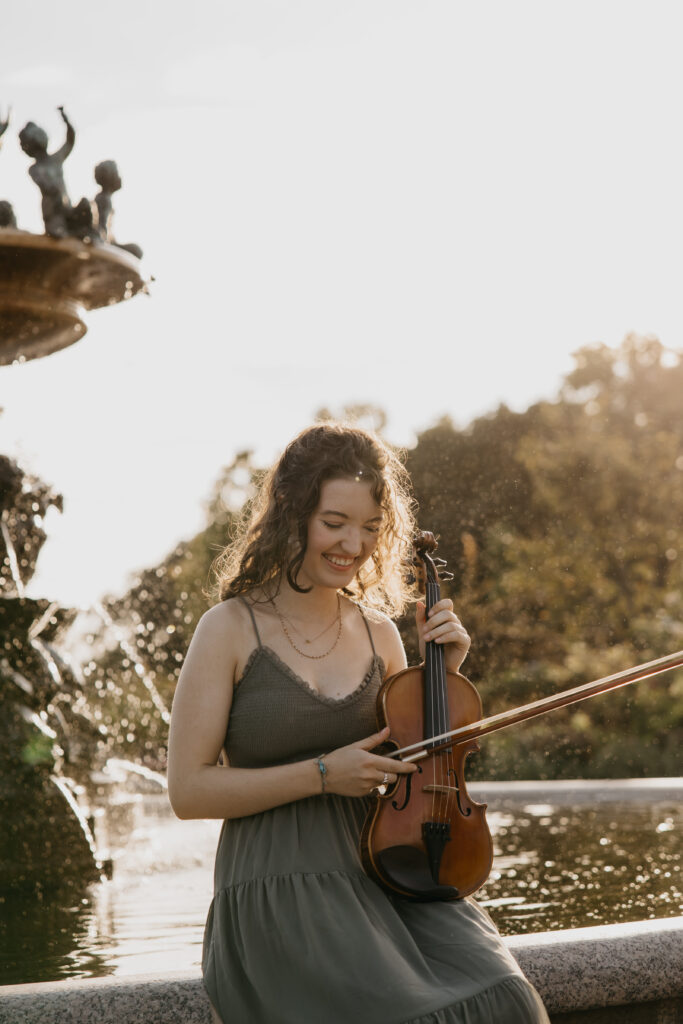 Senior girl in a gray dress playing the violin by a fountain during golden hour, captured in Minneapolis senior photos by Mycah Bain Photography.