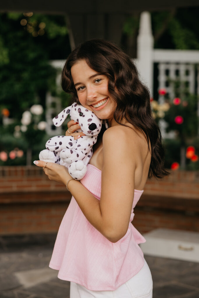 Senior girl in a pink top smiling and hugging her stuffed Dalmatian, photographed outdoors with flowers in the background during her senior portraits by Mycah Bain Photography.