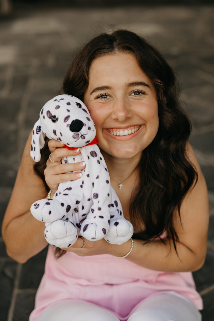 High school senior girl smiling while holding her childhood Dalmatian stuffed animal, wearing a pink top and white pants during senior photos by Mycah Bain Photography in Minneapolis.