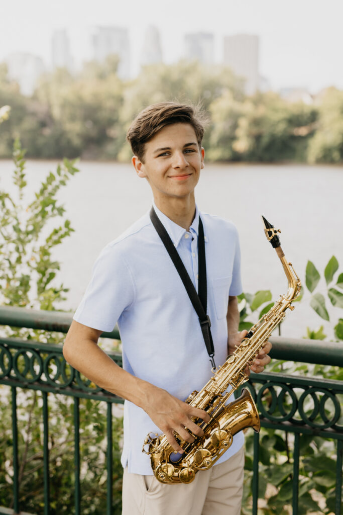 High school senior in a light blue shirt holding his saxophone, standing riverside with downtown Minneapolis in the distance during portraits with Mycah Bain Photography.