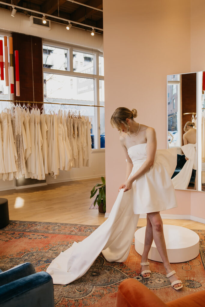 Bride in a strapless mini dress with detachable train admires the fabric during a bridal gown fitting at Vow’d Weddings in Minneapolis.
