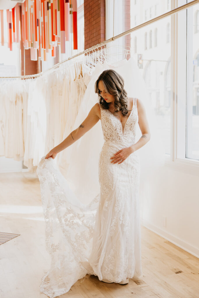 Bride in an elegant lace wedding gown poses by the window, showcasing the gown’s intricate lace details at Vow’d Weddings in Minneapolis.