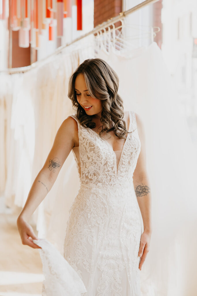 Bride in a sleeveless lace gown with deep V neckline smiles while admiring the dress details at Vow’d Weddings Minneapolis.