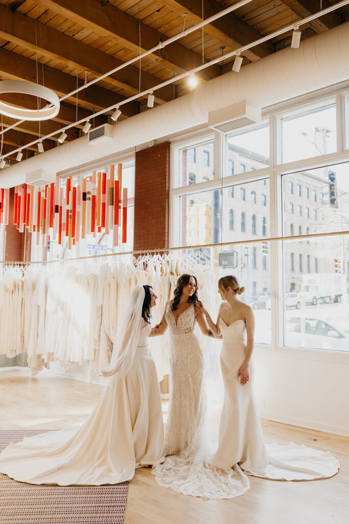 Three brides in elegant gowns share a joyful moment at Vow’d Weddings boutique in Minneapolis, showcasing modern bridal styles.