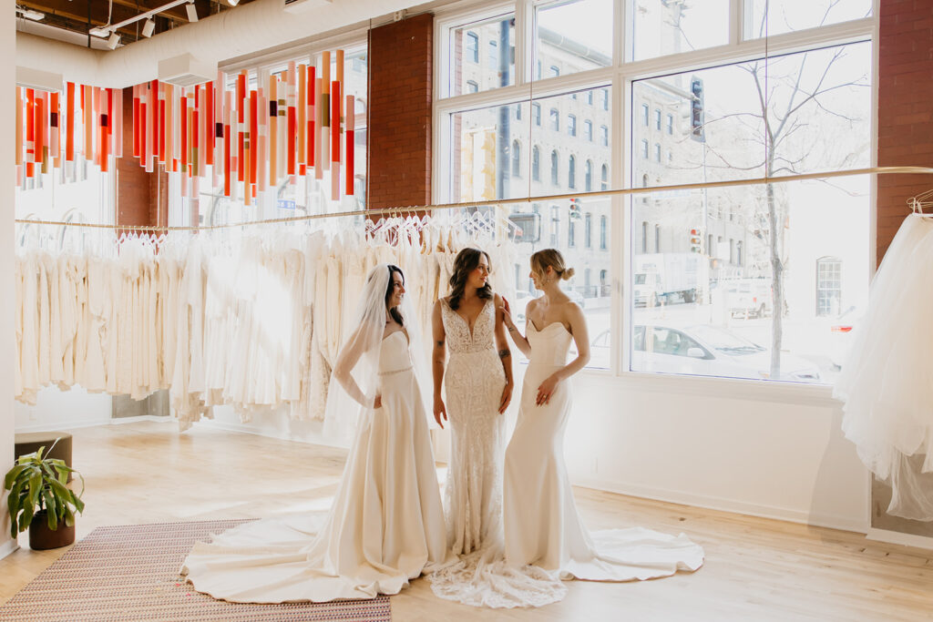 Brides in satin and lace gowns laugh together during a Minneapolis bridal gown photoshoot at Vow’d Weddings.