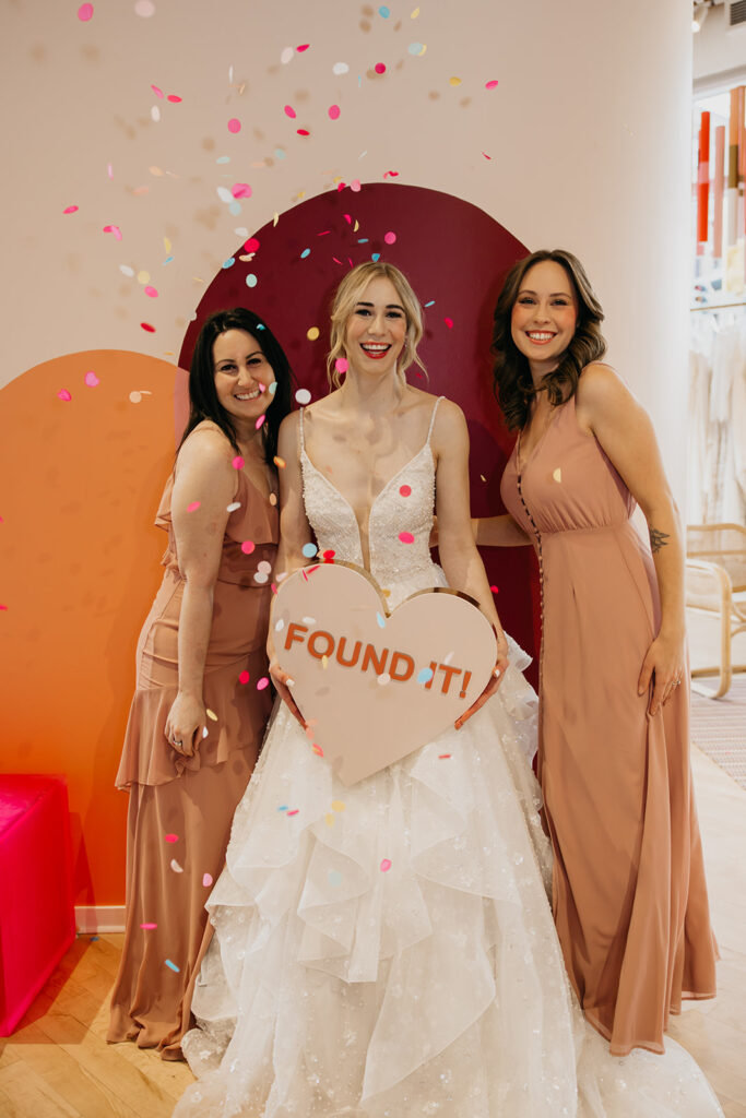 Bride in a sparkling layered gown celebrates with friends at Vow’d Weddings in Minneapolis, holding a heart-shaped “Found It!” sign as confetti falls.