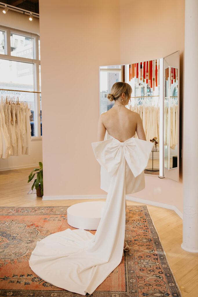 Back view of a bride wearing a short strapless dress with a dramatic oversized bow and detachable train at Vow’d Weddings in Minneapolis.