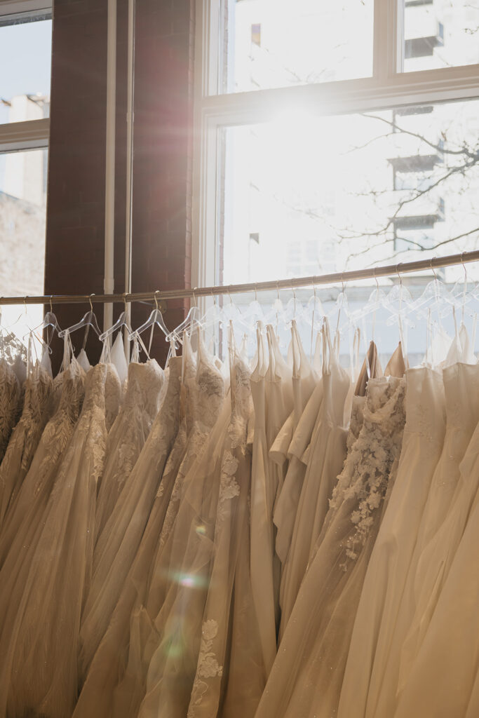 A row of lace and satin bridal gowns hanging at Vow’d Weddings boutique in Minneapolis with natural sunlight streaming through the window.