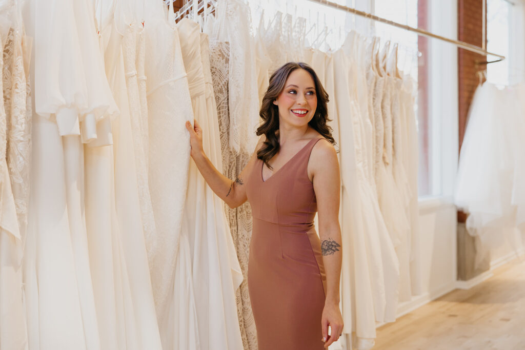Bridesmaid in a fitted mauve dress stands beside racks of lace wedding gowns at Vow’d Weddings in Minneapolis.