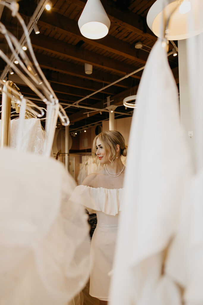 Bride in an off-the-shoulder white dress smiles while browsing wedding gowns at Vow’d Weddings in Minneapolis.