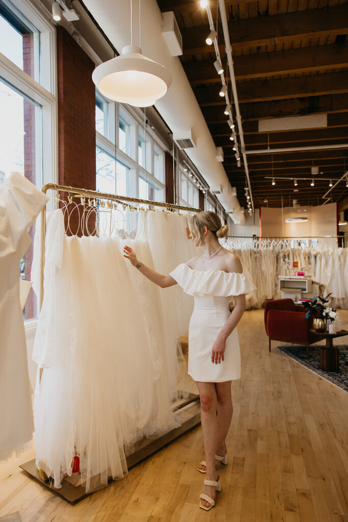 Bride in a short off-the-shoulder dress looks through racks of bridal gowns at Vow’d Weddings boutique in Minneapolis.