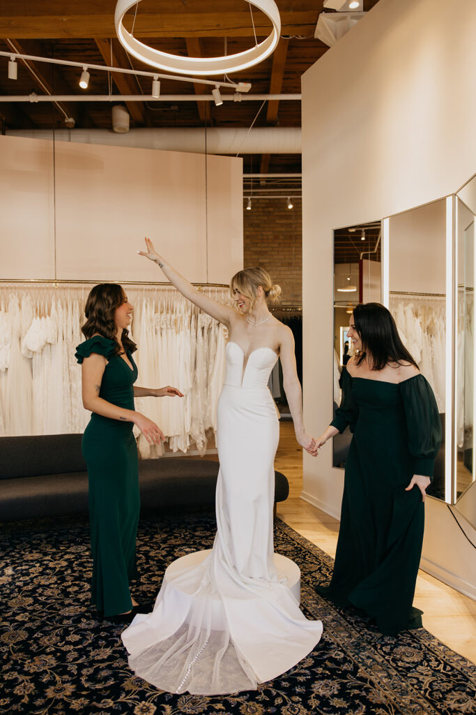 Bride in a sleek strapless gown twirls playfully with bridesmaids during a bridal gown fitting at Vow’d Weddings in Minneapolis.