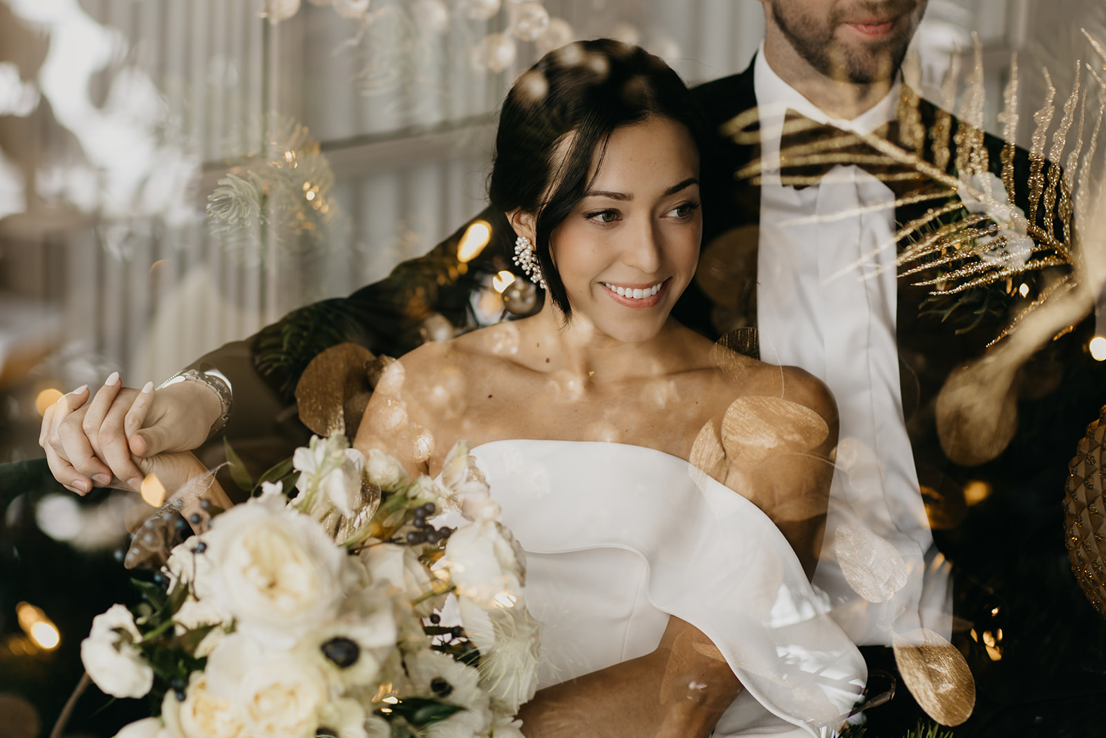 Bride in elegant off-shoulder gown smiling while holding a white floral bouquet, with groom in tuxedo beside her, captured with dreamy golden reflections.