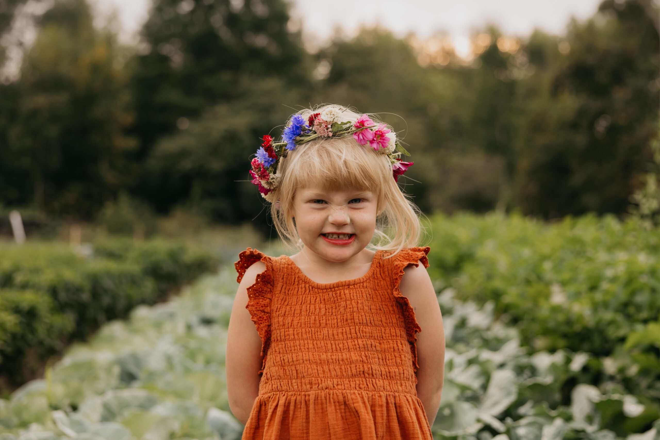 Smiling young girl wearing a rust-colored dress and flower crown in a vegetable field at Abraham’s Table Farm – Minnesota farm brand photography