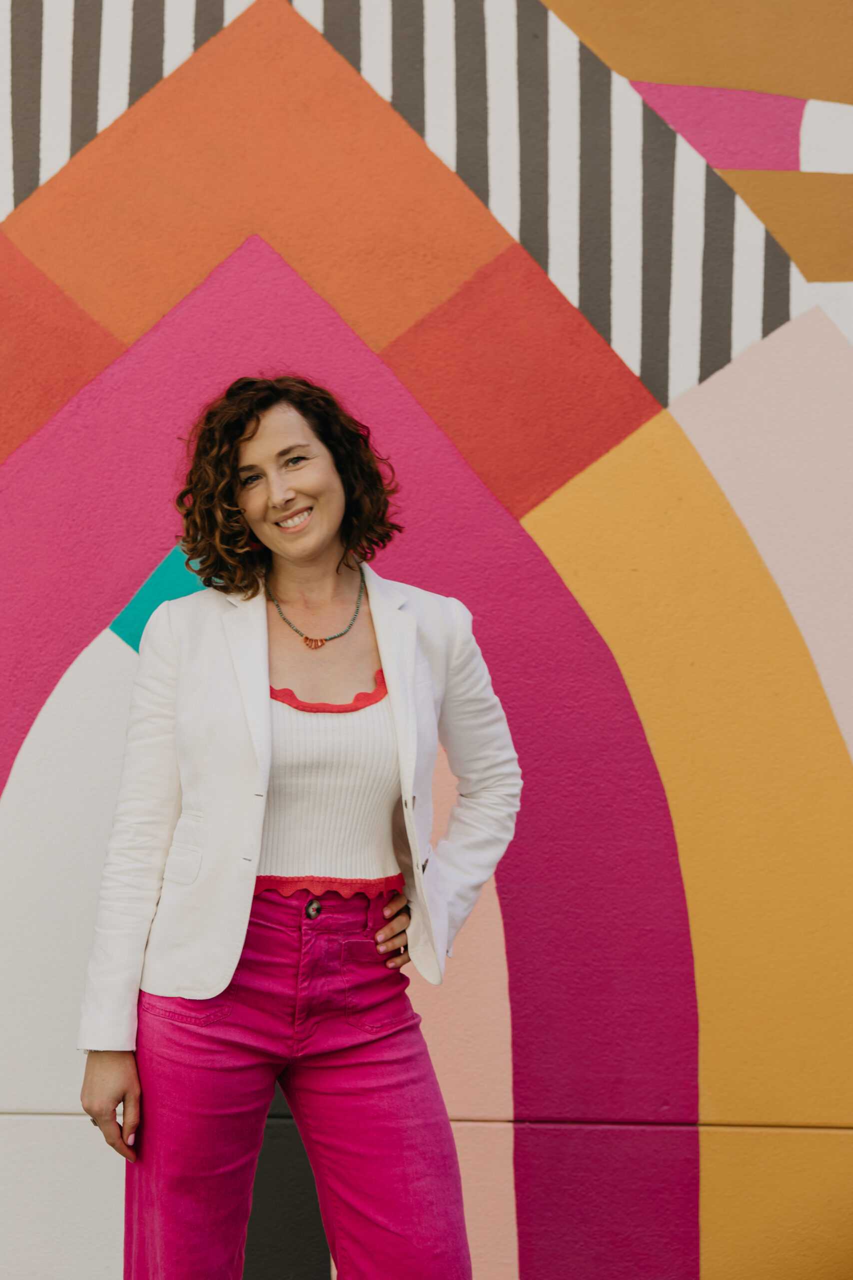 Anne Deanovic, founder of Hot Mustard PR, poses in front of a colorful geometric mural during her Minneapolis brand photoshoot with Mycah Bain Photography. She wears a white blazer, pink pants, and a red-trimmed tank top, representing her creative, bold PR brand.