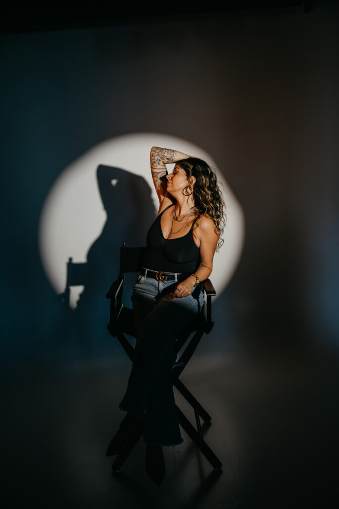 Dramatic studio portrait of Dr. Lauryn in black attire sitting in a director’s chair under a spotlight for her editorial brand session.